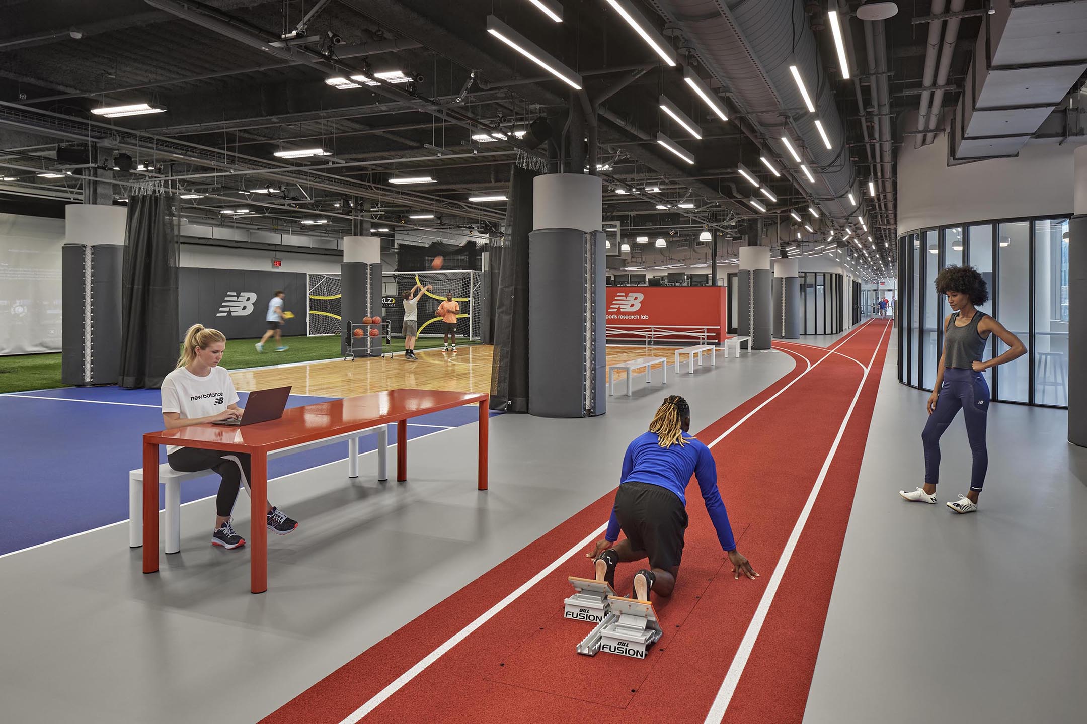 An indoor training area at The TRACK at New Balance, featuring a short red running track, a turf area, and a wooden court. A person is in a starting block on the track, another is at a laptop on a table, and others are in the background.