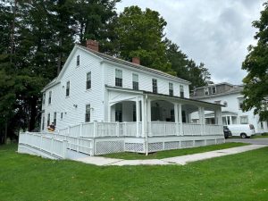 White two-story house with porch and wheelchair ramp, person in safety vest nearby amid trees.