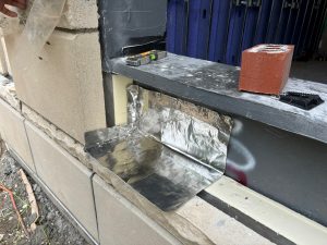 Window sill installation detail showing metal flashing, stone blocks, and insulation, with black frame and temporary brick weight.