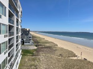 Elevated coastal view showing modern white high-rise with balconies along sandy beach, smaller buildings in distance.