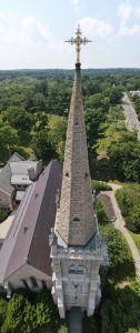 Aerial view of Gothic church spire with golden cross, rising above cemetery and wooded neighborhood.